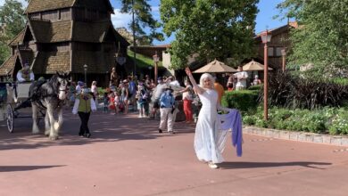 A Disney cast member in a white dress waves, guiding a horse-drawn carriage and guests by themed buildings on a wintry Main Street.