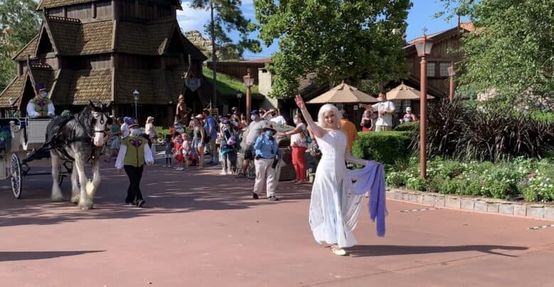 A Disney cast member in a white dress waves, guiding a horse-drawn carriage and guests by themed buildings on a wintry Main Street.
