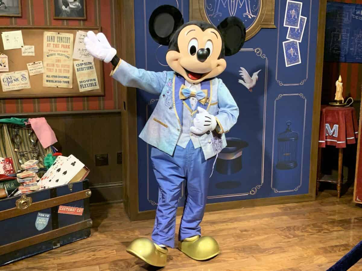 Mickey Mouse greets guests in a blue suit inside a magical Disney World room, surrounded by themed props and ticket offer signs.