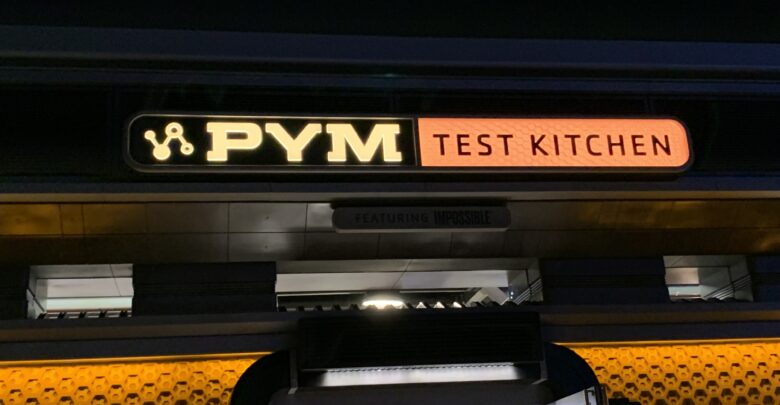 PYM TEST KITCHEN sign glows in yellow and orange above the Avengers Campus entrance at night, inviting guests inside.