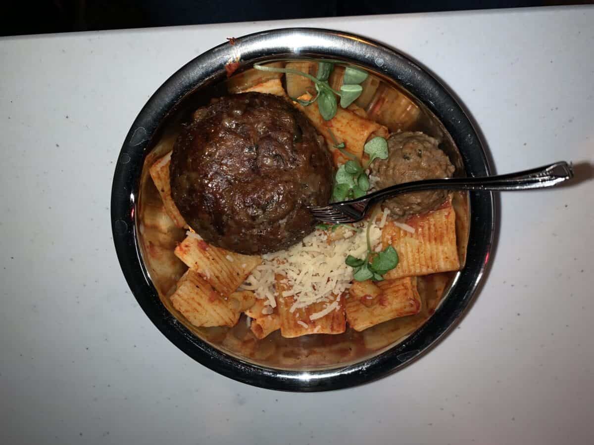 Super-sized meatball atop pasta with cheese and greens at Avengers Campus, served in a metal bowl with a side fork.