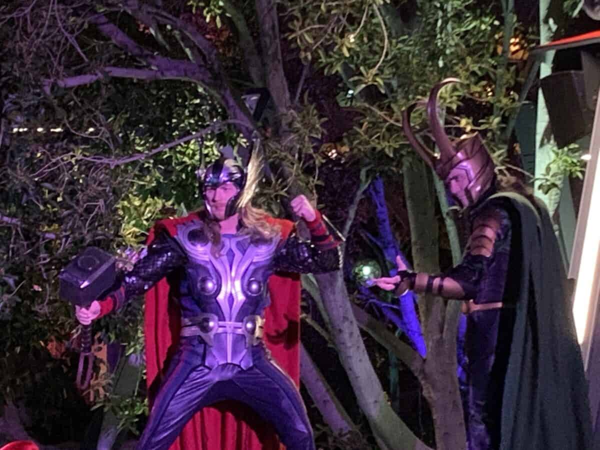 Thor and Loki strike dynamic poses at Avengers Campus, with glowing nighttime lights and lush trees setting the heroic scene.