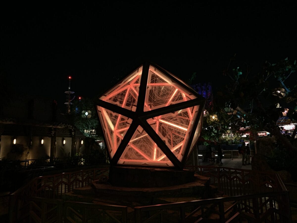 A glowing geometric sculpture with vibrant orange lights brightens Avengers Campus at night, encircled by a themed safety fence.