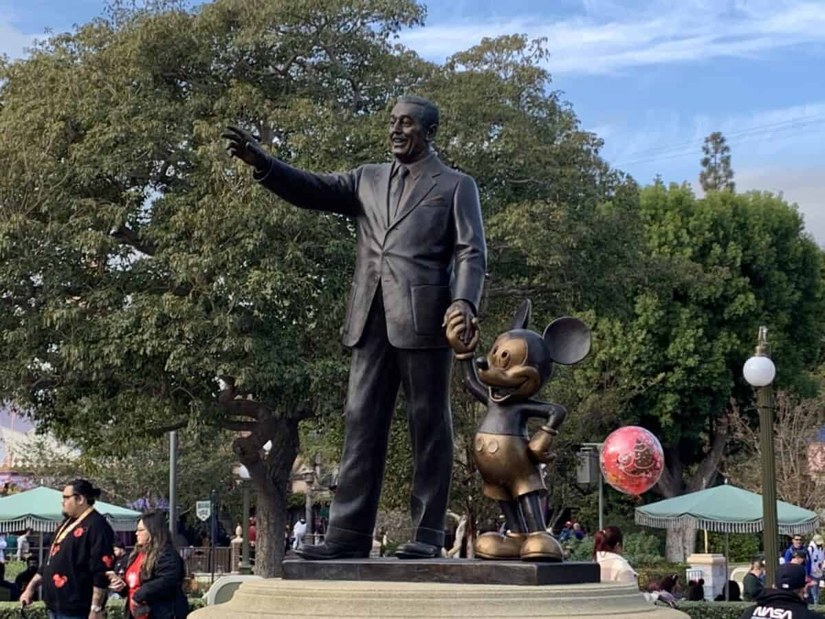 Walt Disney and Mickey Mouse statue in Magic Kingdom park, with guests and Disney Park Pass Reservations in the background.