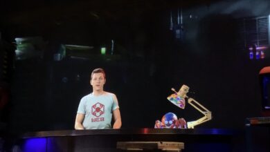 A robotics engineer in a WEB t-shirt showcases a mini bot and vibrant gear at Innovators Lab, lit by moody show lighting.