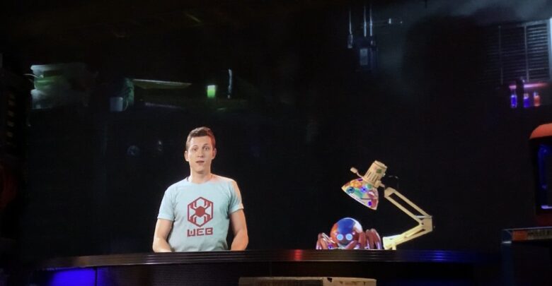 A robotics engineer in a WEB t-shirt showcases a mini bot and vibrant gear at Innovators Lab, lit by moody show lighting.