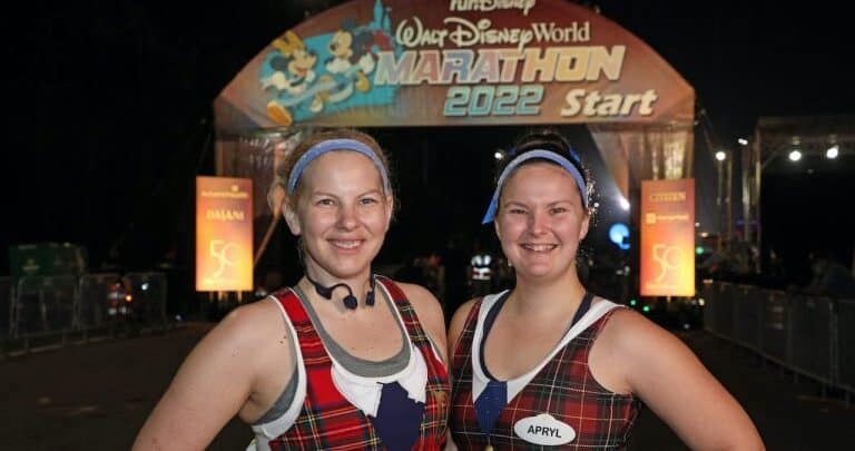 Two runners in plaid gear beam at the 2022 Walt Disney World Marathon start line, Cinderella Castle glowing behind them at night.