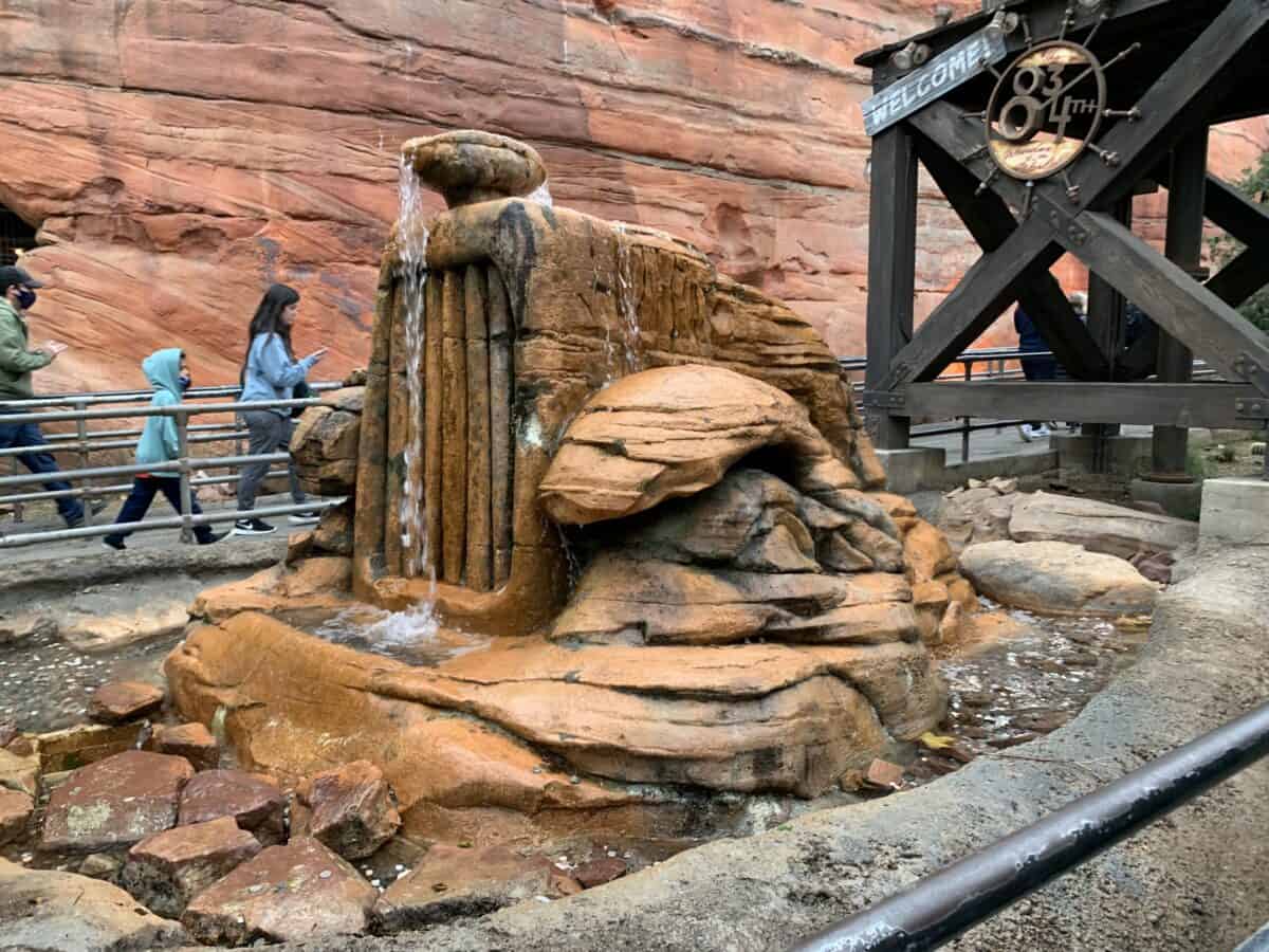 Rust-hued rock fountain with cascading water, guests strolling by, and a classic wooden Disneyland sign setting the scene.