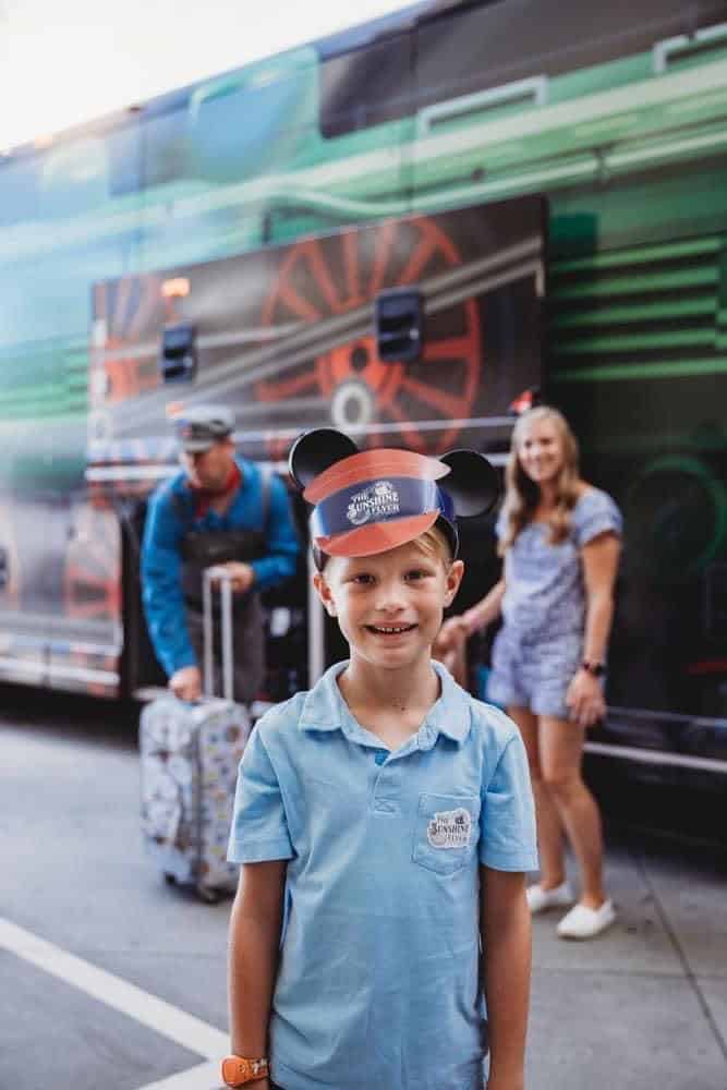 Young Disney fan in a conductor hat and Mickey ears beams by the Sunshine Flyer bus at Orlando Airport, magic trip about to begin.