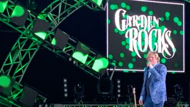 A Garden Rocks musician in a blue jacket rocks the stage under green lights, with the Garden Rocks sign glowing behind them.