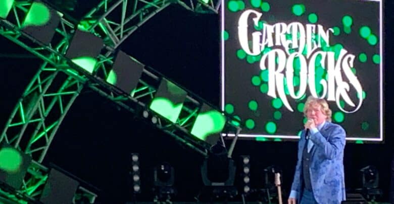 A Garden Rocks musician in a blue jacket rocks the stage under green lights, with the Garden Rocks sign glowing behind them.