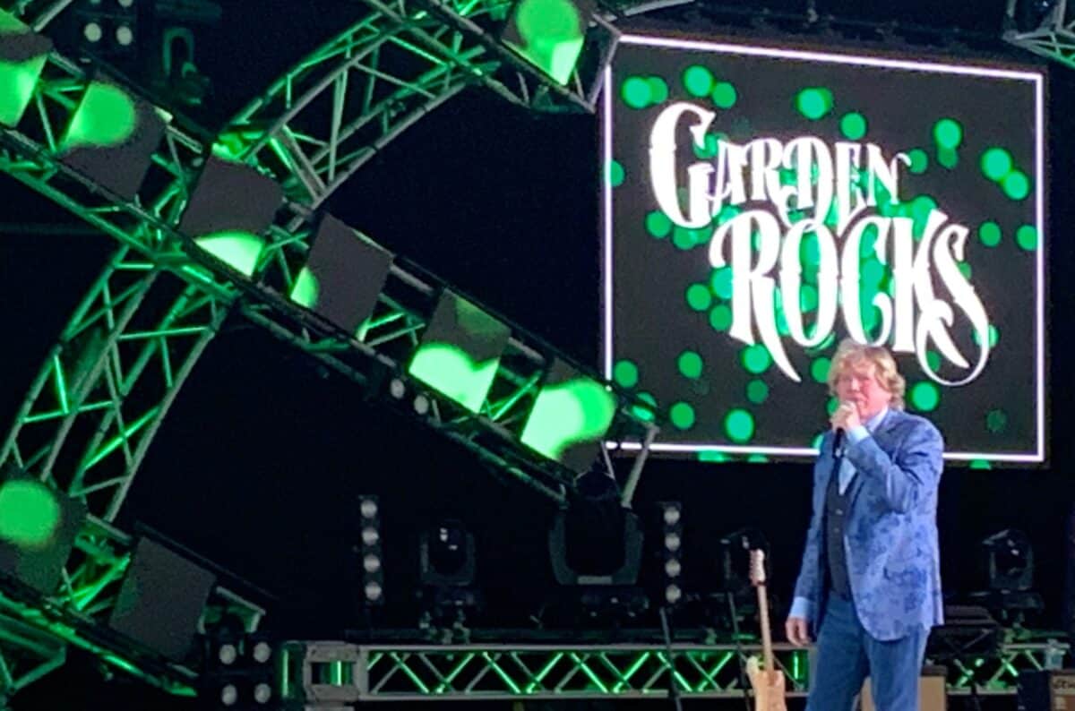 A Garden Rocks musician in a blue jacket rocks the stage under green lights, with the Garden Rocks sign glowing behind them.