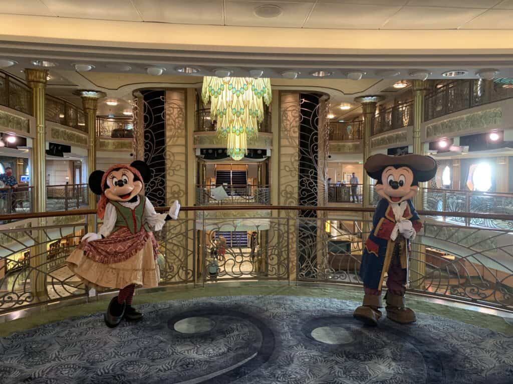 Pirate Minnie and Mickey Mouse pose on the Disney Fantasy’s grand balcony beneath a striking green chandelier, ready for fun.