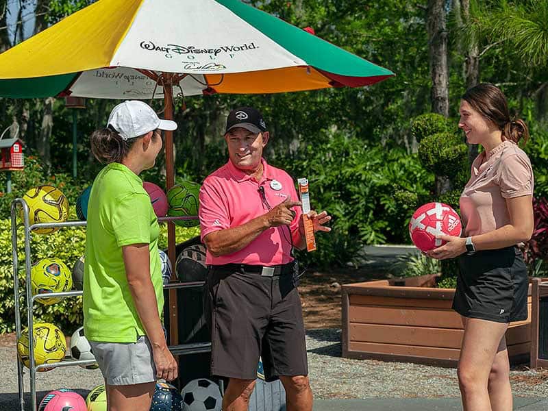 Guests laugh and chat by colorful soccer balls beneath a Disney Parks umbrella, exploring FootGolf fun at the resort.