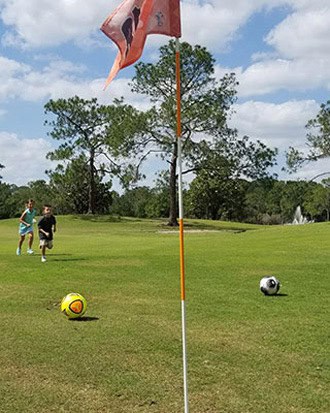 Kids dash across a green fairway toward a footgolf flag, soccer balls ahead, under clear skies—FootGolf fun for all ages!.