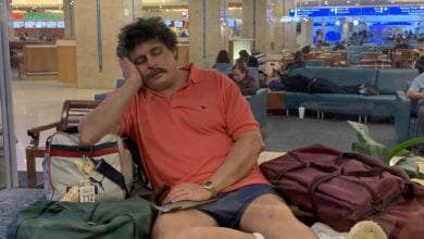 A traveler in a red polo shirt naps near his luggage in the airport, catching some rest before flying to the theme park merger event.