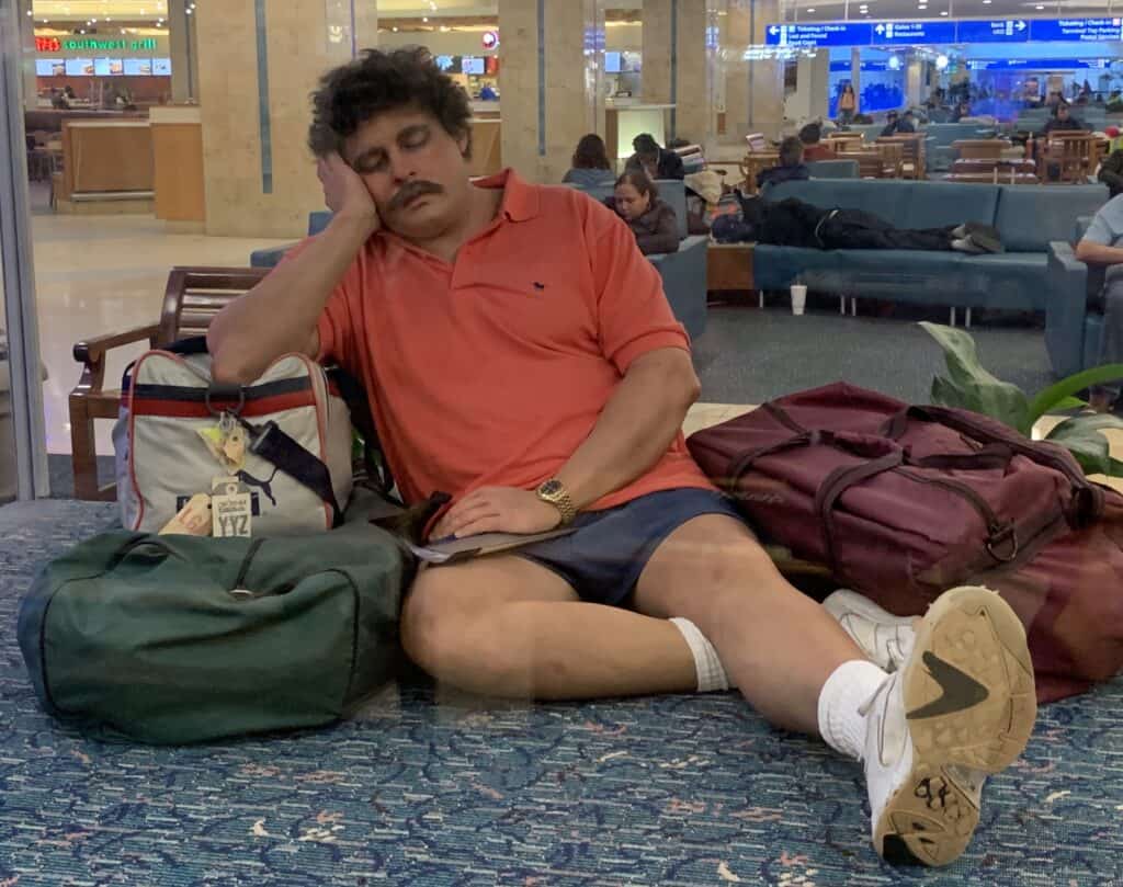 A guest in casual attire naps on the airport floor with bags nearby, likely awaiting delayed flights as airline merger news circulates.