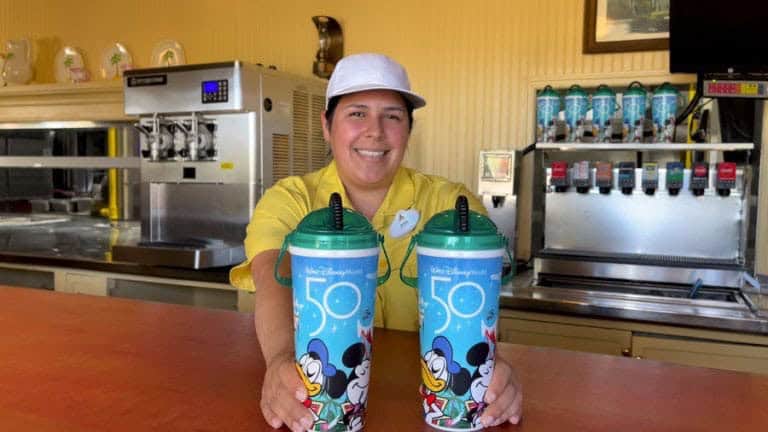 Cast member in a yellow shirt serves drinks with two Mickey Mouse “50” souvenir cups at Disney’s themed beverage station.