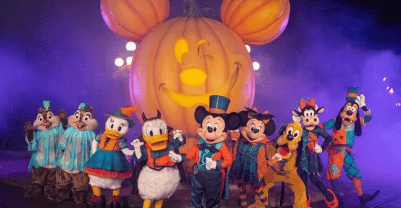 A group of Disney characters, including Mickey Mouse, Donald Duck, Goofy, and Pluto, dressed in Halloween costumes, stand in front of a large illuminated pumpkin with Mickey Mouse's face carved into it. The background is filled with purple mist.