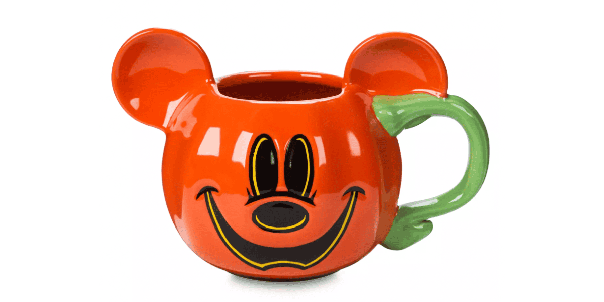 A colorful mug in the shape of a character's head, featuring a bright orange color with a wide, joyful smile and black eyes. The mug has two large ears and a large green handle shaped like a twisted vine.