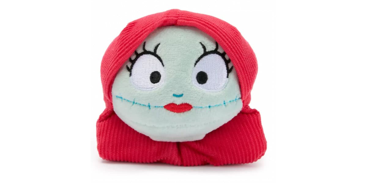 A plush toy with a pale green face, large embroidered eyes, red lips, black stitched smile, and bright red hair styled in two sections, resembling Sally from "The Nightmare Before Christmas.
