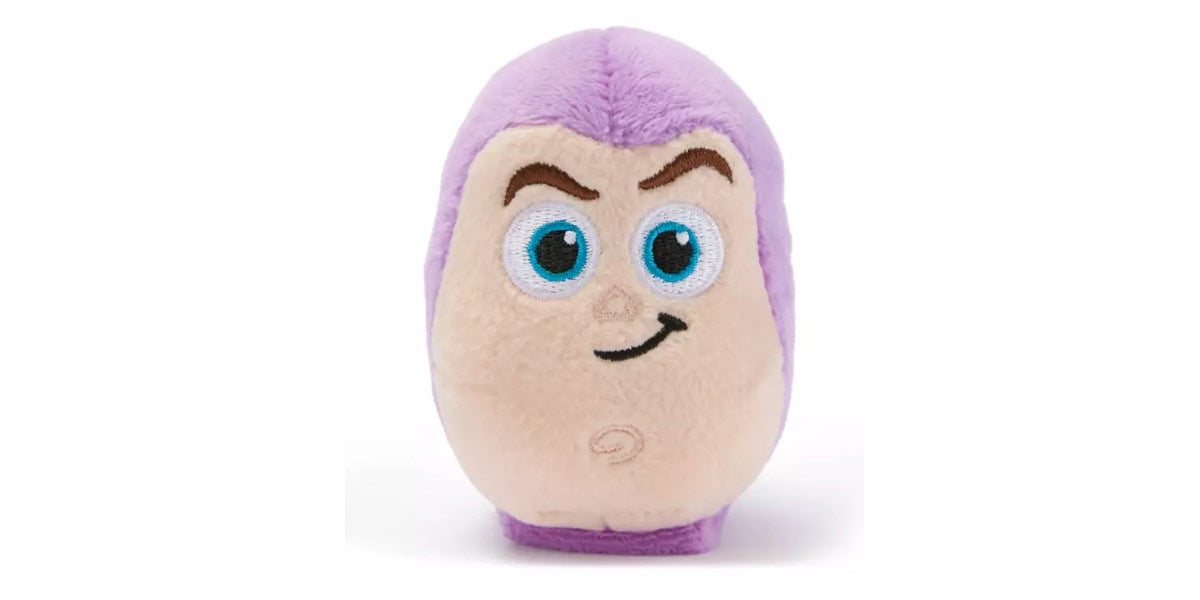 A small, egg-shaped plush toy with a light brown face, large blue eyes, dark eyebrows, a playful smirk, and purple fabric on the top and sides of its head.