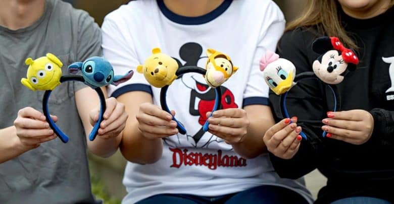 Three people holding Disney-themed headbands with plush characters: Alien, Stitch, Winnie the Pooh, Tigger, Daisy Duck, and Minnie Mouse. The person in the center wears a white Disneyland shirt.