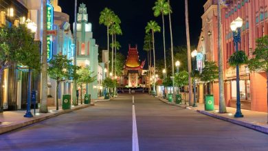 A brightly lit, empty street at night with colorful art deco buildings, palm trees, and the replica of the Chinese Theatre in the distance, reminiscent of a Hollywood-themed park.