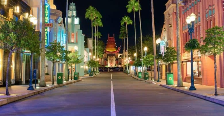 A brightly lit, empty street at night with colorful art deco buildings, palm trees, and the replica of the Chinese Theatre in the distance, reminiscent of a Hollywood-themed park.