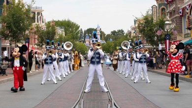 A marching band performs on a street with brass instruments, flanked by people in Mickey and Minnie Mouse costumes. Colorful buildings and trees line the street, while talk swirls—could Disney World follow Disneyland and remove early park entry? A castle rises in the background.