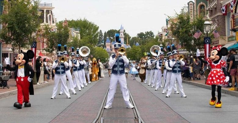 A marching band performs on a street with brass instruments, flanked by people in Mickey and Minnie Mouse costumes. Colorful buildings and trees line the street, while talk swirls—could Disney World follow Disneyland and remove early park entry? A castle rises in the background.