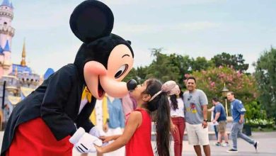 A girl in a red dress happily kisses Mickey Mouse on the nose at Disney, with other visitors and the castle blurred in the background with a Disney World annual pass guest.