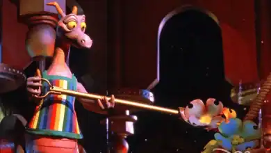 A purple dragon wearing a striped apron holds a long-handled spoon over a flower-shaped bowl in a whimsical, colorful setting.