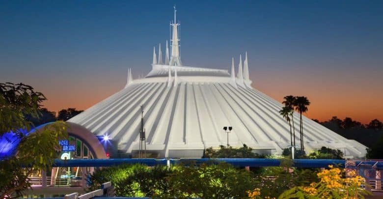 A futuristic white, conical building with spires, surrounded by greenery, is lit against a colorful sunset sky. This is Space Mountain, an amusement ride at a theme park.