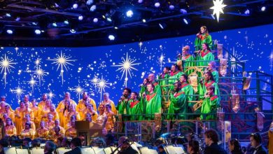 A large choir in yellow and green robes sings on a tiered stage decorated with bright star lights, backed by an orchestra. The backdrop is blue with glowing stars, creating a festive, celebratory atmosphere.