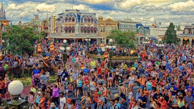 A large, colorful crowd fills a lively theme park street lined with vibrant, ornate buildings. Halloween decorations and pumpkins are visible, and the sky is partly cloudy, adding to the festive atmosphere.