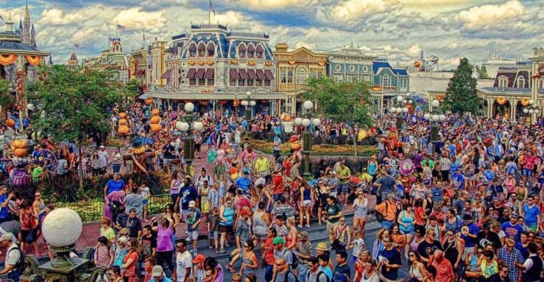 A large, colorful crowd fills a lively theme park street lined with vibrant, ornate buildings. Halloween decorations and pumpkins are visible, and the sky is partly cloudy, adding to the festive atmosphere.
