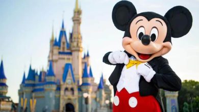 Mickey Mouse, wearing his classic outfit with red shorts, yellow bow tie, and white gloves, poses cheerfully in front of a blue and gold castle at a Disney theme park.