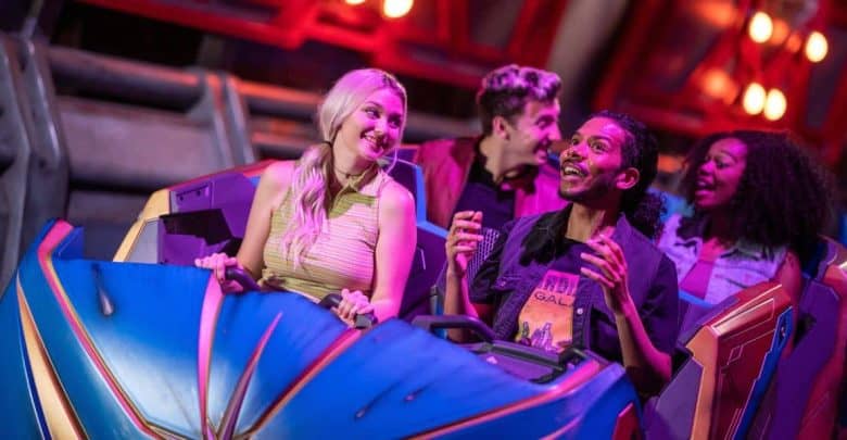 Four people smiling and laughing as they ride a colorful amusement park roller coaster, with bright lights and metallic structures in the background.