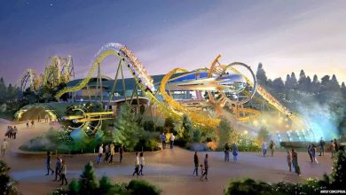 Artist’s illustration of a vibrant amusement park at dusk, featuring a twisting yellow and green roller coaster with people riding, surrounded by trees, glowing lights, and visitors walking below.