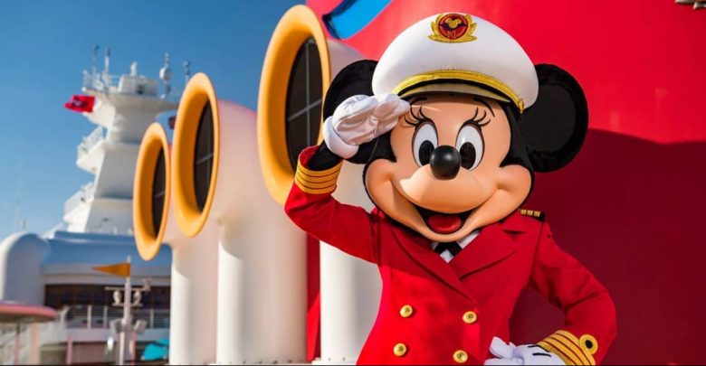 Minnie Mouse, dressed as a ship captain in a red uniform and white hat, stands and salutes in front of large yellow and red cruise ship funnels.