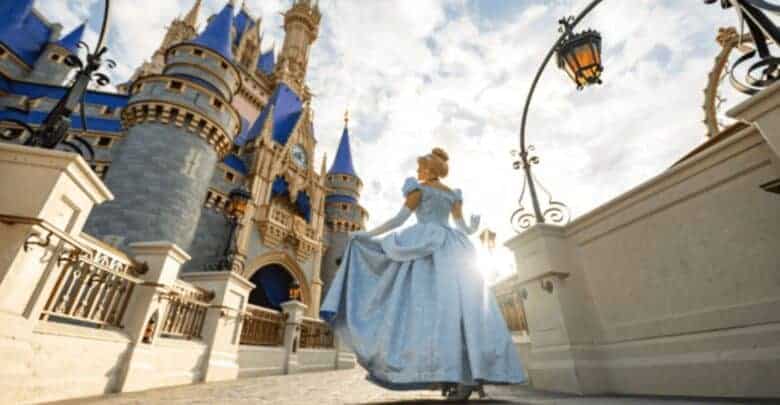 A woman in a blue ball gown walks toward a fairytale castle with blue spires under a partly cloudy sky, sunlight shining behind her. Ornate street lamps and castle details frame the scene.