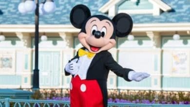 A person in a Mickey Mouse costume is standing outdoors in front of a pastel-colored building with blue trim, extending their right arm. Mickey is dressed in his signature red shorts with white buttons, yellow bow tie, and black jacket at a Disney Park Resort in Tokyo for summer 2025.