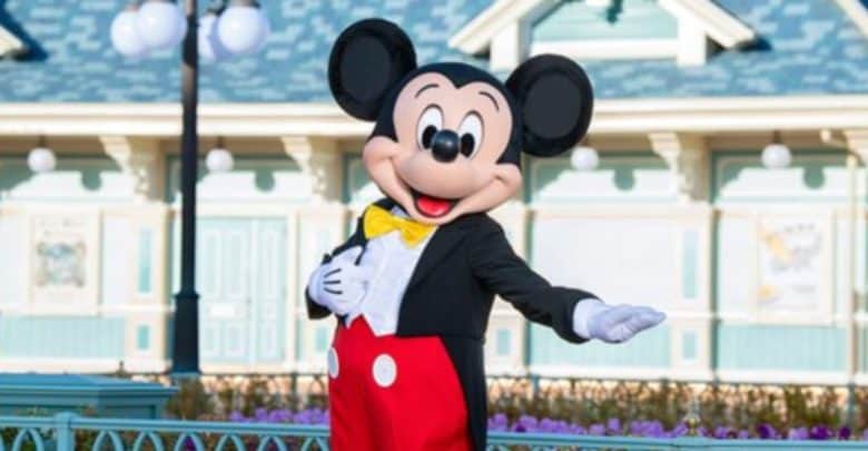 A person in a Mickey Mouse costume is standing outdoors in front of a pastel-colored building with blue trim, extending their right arm. Mickey is dressed in his signature red shorts with white buttons, yellow bow tie, and black jacket at a Disney Park Resort in Tokyo for summer 2025.