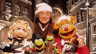 A man in a nightcap stands smiling with Muppet characters, including Miss Piggy, Fozzie Bear, Kermit the Frog, and others, in a snowy, old-fashioned street setting.