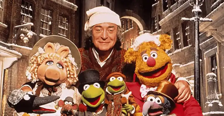 A man in a nightcap stands smiling with Muppet characters, including Miss Piggy, Fozzie Bear, Kermit the Frog, and others, in a snowy, old-fashioned street setting.