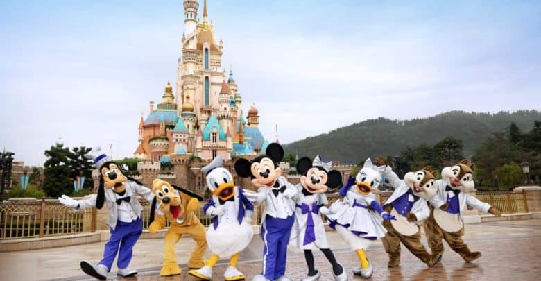 Disney characters Goofy, Pluto, Donald Duck, Mickey Mouse, Minnie Mouse, Daisy Duck, Chip, and Dale pose happily in front of a colorful castle at a Disney theme park, with a clear sky and greenery in the background.