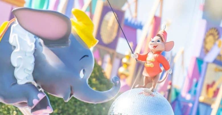 A Dumbo elephant statue with a yellow hat faces a smiling mouse figure in a red uniform standing on a reflective silver ball. Colorful decorations and geometric patterns are visible in the blurred background.