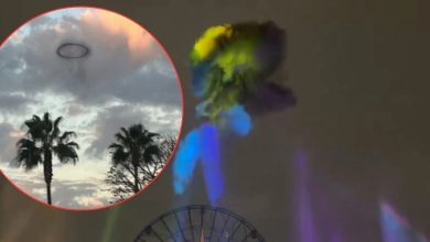 A dark ring-shaped cloud floating above palm trees and a ferris wheel in a cloudy sky, with colorful lights beaming upward.