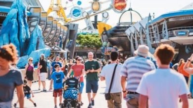 People walk through a busy, colorful theme park area with futuristic decor and attractions. Shops and rides line the path, and various families and individuals can be seen, some with strollers and others taking photos. The atmosphere is vibrant and lively.